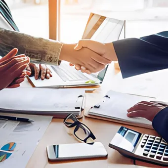 Investitori che si stringono la mano su una scrivania con documenti, calcolatrice e laptop